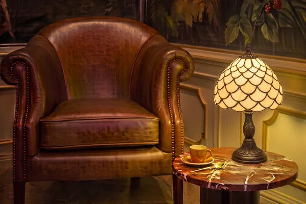 Brandende Tiffany tafellamp met geschubd crème-amber glaspatroon op een bijzettafel naast een leren fauteuil in een klassieke leeshoek | RORUVERA