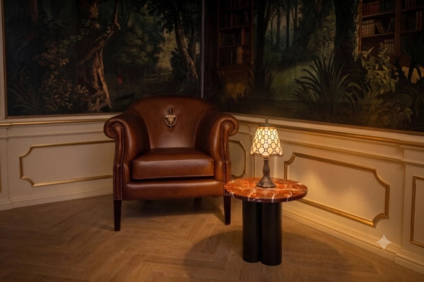 Brandende Tiffany tafellamp 7004 met crème-amber glas en ronde sieraccenten op een marmeren tafel naast een leren fauteuil  RORUVERA