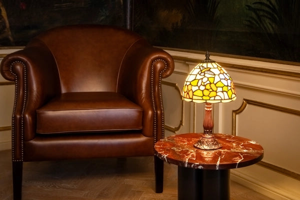 7005 - Tiffany tafellamp met kolibrie- en bloemmotief op een marmeren bijzettafel naast een leren fauteuil | RORUVERA