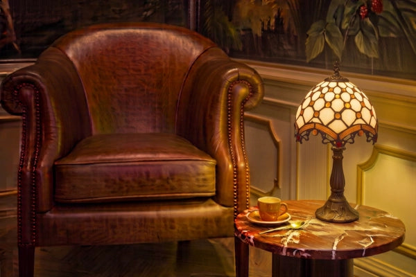 Tiffany tafellamp met crème glas-in-lood kap op een rond bijzettafeltje naast een klassieke leren fauteuil in een warme leeshoek | RORUVERA