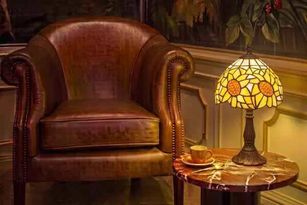 Brandende Tiffany tafellamp 7007 met geel-oranje zonnebloemmotief op een marmeren bijzettafel in een klassieke leeshoek | RORUVERA