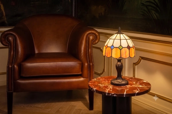 Brandende Tiffany tafellamp 7010 met crème-amber glas en gekleurde ruitaccenten op een marmeren tafel naast een leren fauteuil | RORUVERA