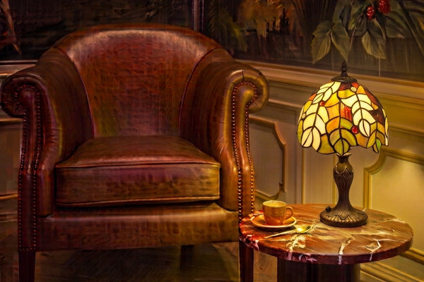 Tiffany tafellamp met groen en crème bladmotief en warme rode accenten op marmeren bijzettafel naast bruine leren fauteuil | RORUVERA