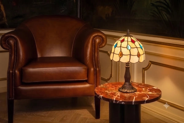 Tiffany tafellamp met crème glas en kleurrijke druppelaccenten op marmeren bijzettafel naast bruine leren fauteuil | RORUVERA