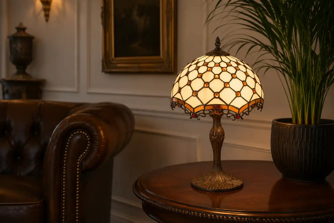 Tiffany tafellamp met crème glas, amberkleurige accenten en sierlijke donkere voet op een houten bijzettafel in een klassieke woonkamer met leren fauteuil en grote groene plant |RORUVERA