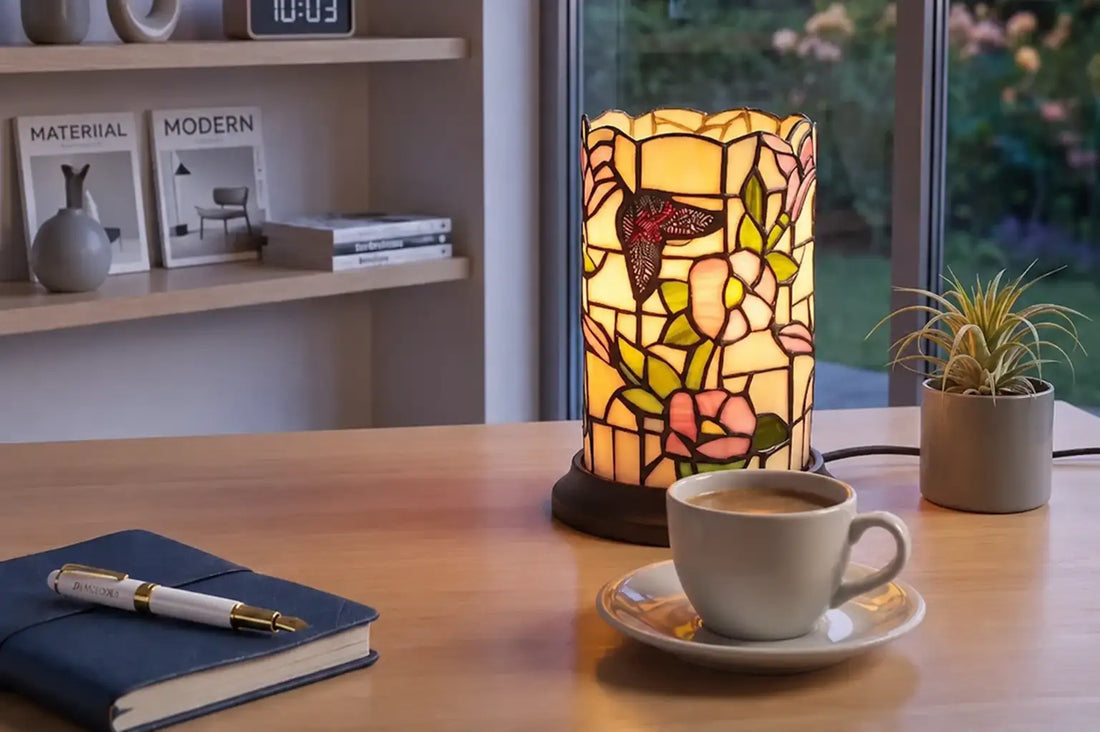 Ingeschakelde Tiffany tafellamp 7175 met vlinder- en bloemenmotief in pastelglas op een modern houten bureau met koffie, notitieboek en witte RORUVERA-vulpen