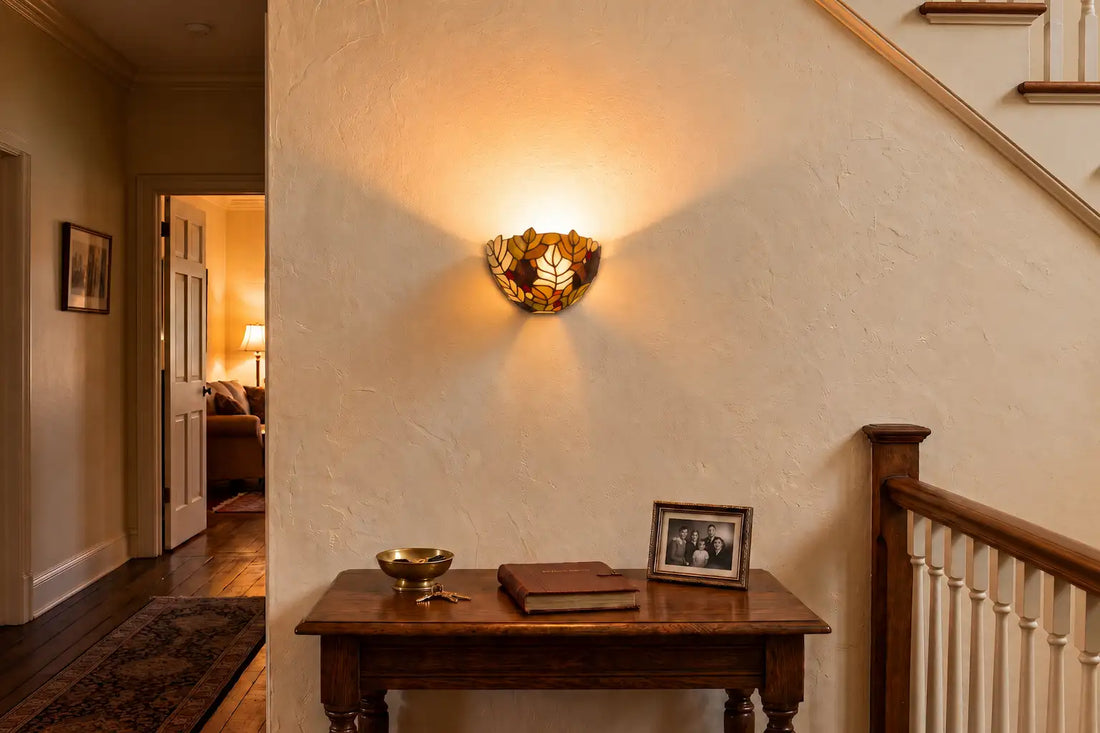 Tiffany wandlamp met bladerenmotief aan een lichte muur boven een houten sidetable in een klassieke hal met trap | RORUVERA